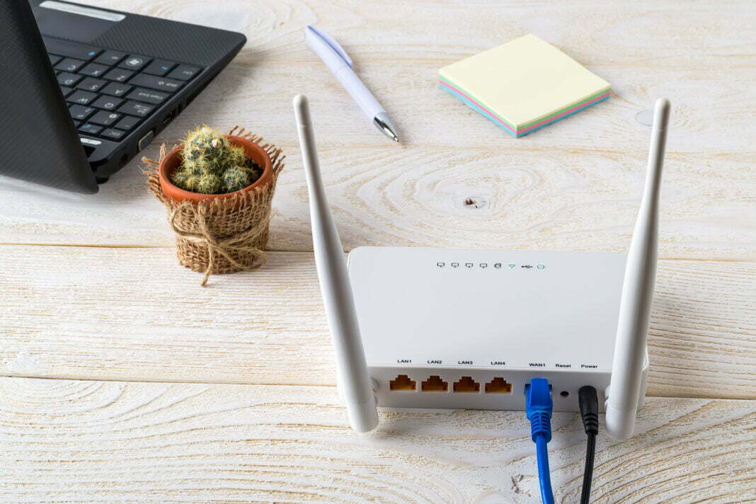 White Wi-Fi wireless router near laptop on a white wooden table. Wlan router with internet cables plugged in on a table in a home or office