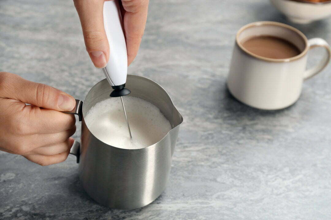 Woman using milk frother in pitcher near cup of coffee on table